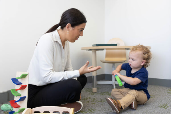 woman kneels on the floor, gesturing with her hand, while a young child sits nearby