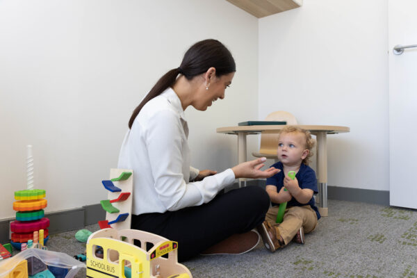 A woman sits on the floor in a playroom, engaging with a toddler