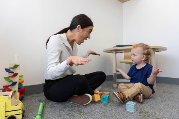 A woman and a young child sit on the floor amidst toys, engaging in an expressive conversation