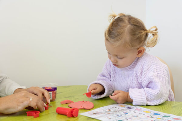 A young child with blonde hair in two pigtails is playing with red play dough at a green table,