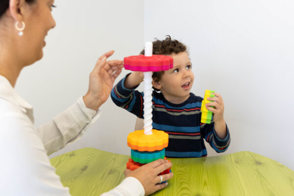 A child and an adult are stacking colorful plastic pieces on a vertical rod at a green table.