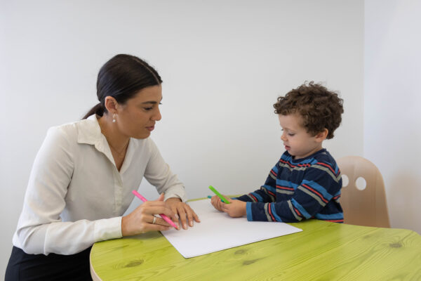 A woman and a child sit at a green table. Woman holds a pink marker, child holds a green marker